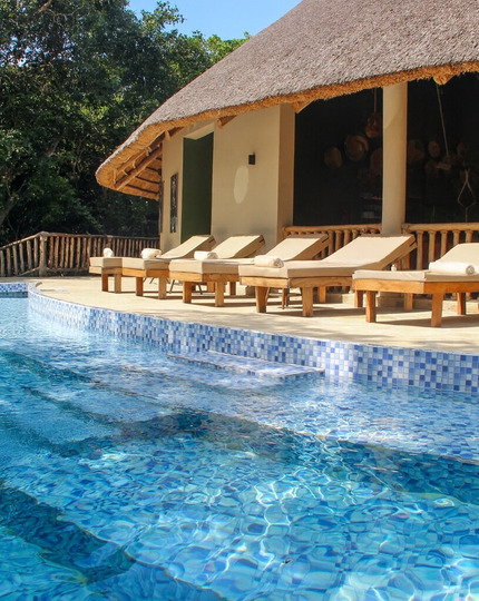 Resort pool with blue mosaic tiles, wooden deck and cushioned sunbeds beneath a thatched terrace by trees.