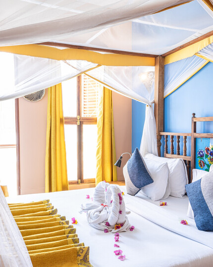 Four‑poster canopy bed with white netting, carved wooden headboard, blue accent wall, yellow curtains, towel swans and flower petals on white linens.
