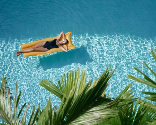 Guest in black swimsuit relaxing on yellow float in clear blue hotel pool surrounded by tropical palm leaves