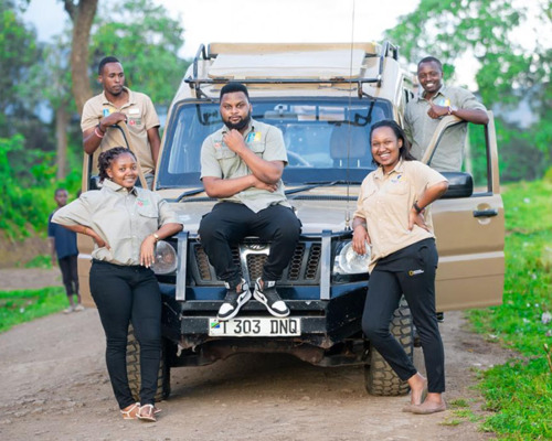 Five safari guides in khaki uniforms posing by a rugged safari vehicle on a dirt road surrounded by greenery