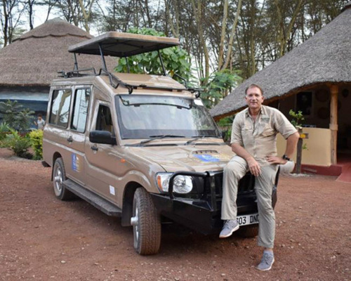 Safari guide in khaki outfit sitting on front of a tan open-roof safari vehicle with thatched-roof lodges and trees in background