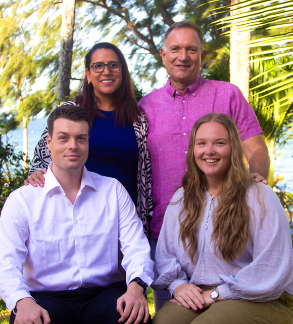 Four smiling guests posing in a shaded beachfront resort garden, palm trees and ocean visible behind them.