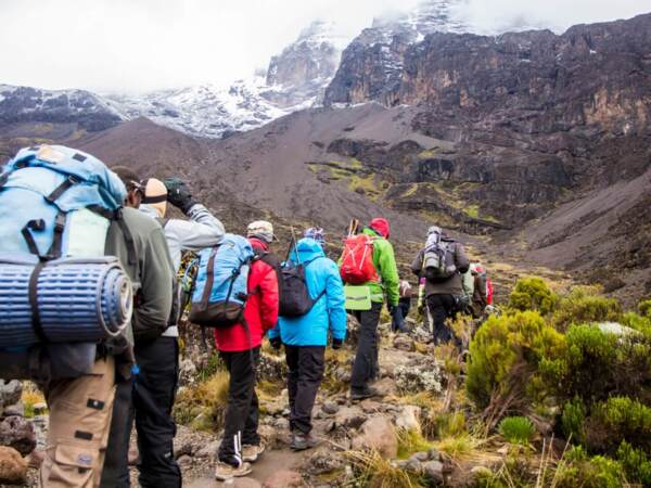 Group of hikers with backpacks trekking on a rocky mountain trail near snow-capped peaks under a cloudy sky