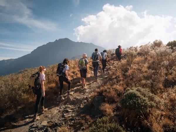 Group of hikers walking up rocky mountain trail surrounded by dry shrubs under a partly cloudy sky