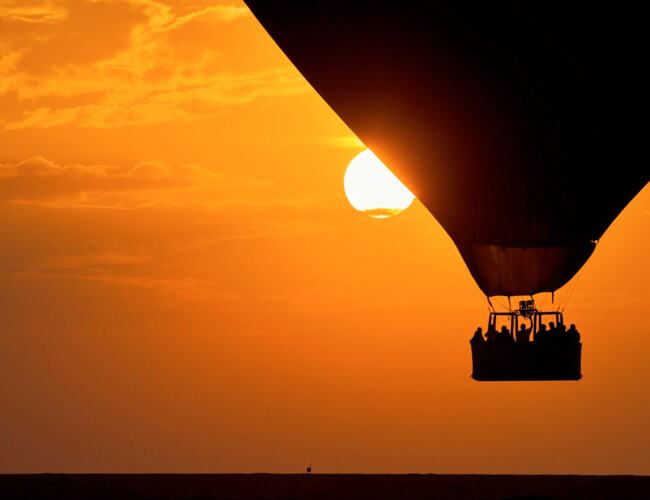Guests enjoy a peaceful hot air balloon ride at sunset with a vivid orange sky and sun partially hidden behind the balloon.