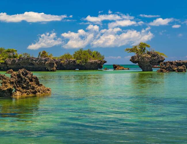 Turquoise shallow coastal water with unique rock formations topped with greenery under a bright blue sky with scattered clouds