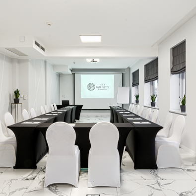 Modern hotel conference room with white chairs, black tablecloths, projector screen, and plants on windowsills.