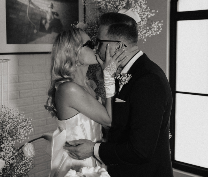 Para młoda całująca się przy obsypanym kwiatami torcie weselnym w eleganckim, przytulnym wnętrzu.