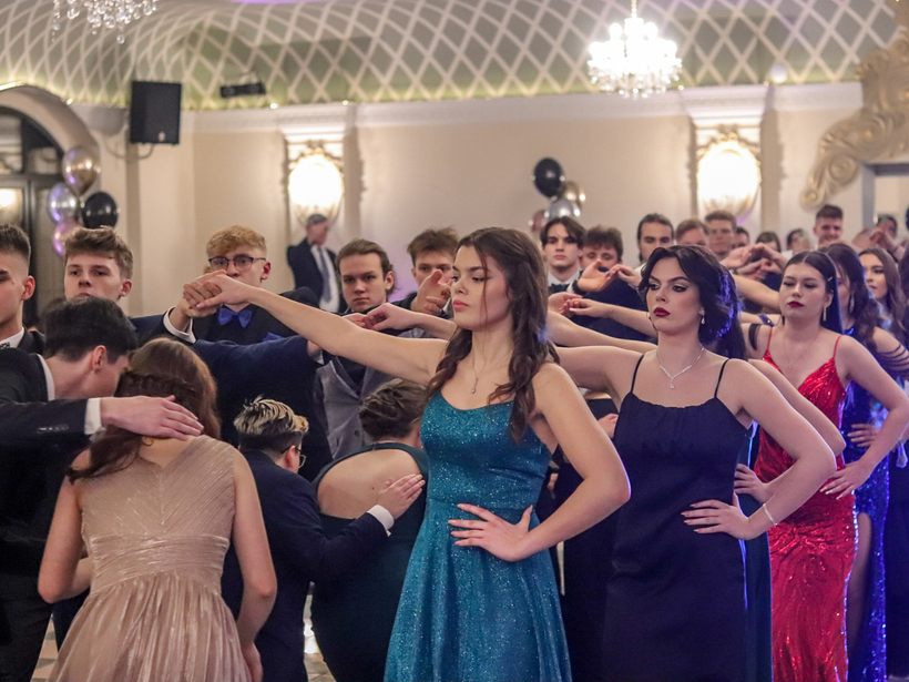 Młodzi goście w eleganckich strojach uczestniczą w formalnym tańcu w stylowej sali balowej z kryształowym żyrandolem.