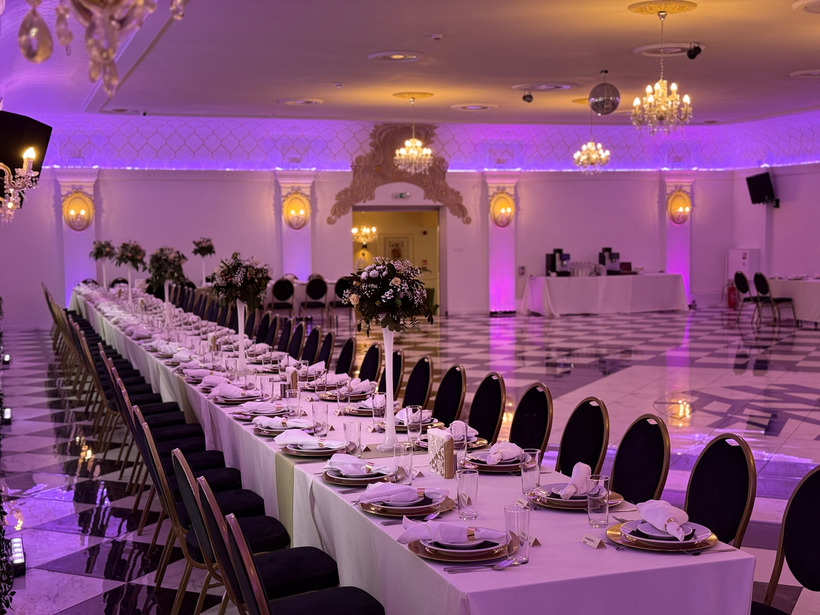 Elegancka sala bankietowa w hotelu ustawiona na przyjęcie — długie stoły, kwiatowe dekoracje, żyrandole, fioletowe oświetlenie i marmurowa podłoga.