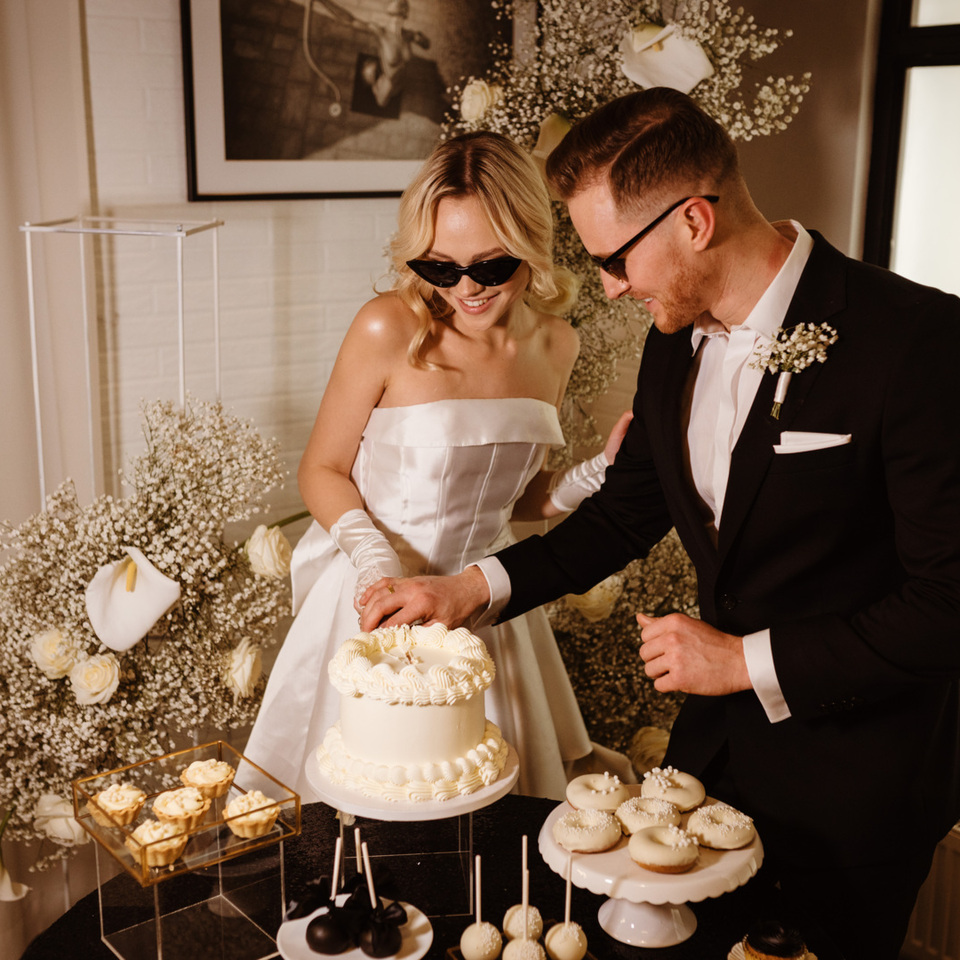 Bride and groom cutting cream wedding cake in a stylish room decorated with flowers and sweet treats.
