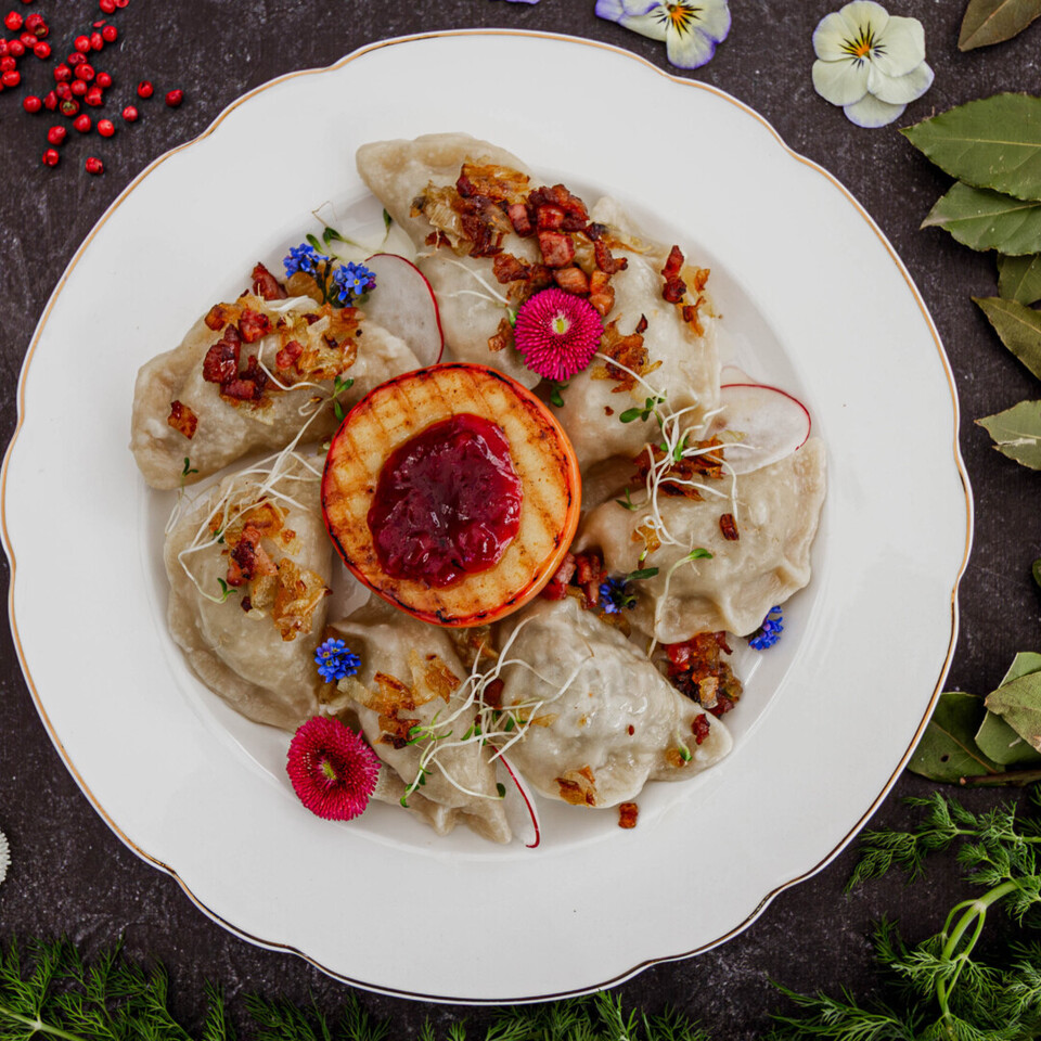 Plate of pierogi topped with sautéed onions, bacon, cranberry sauce, garnished with edible flowers and fresh herbs.