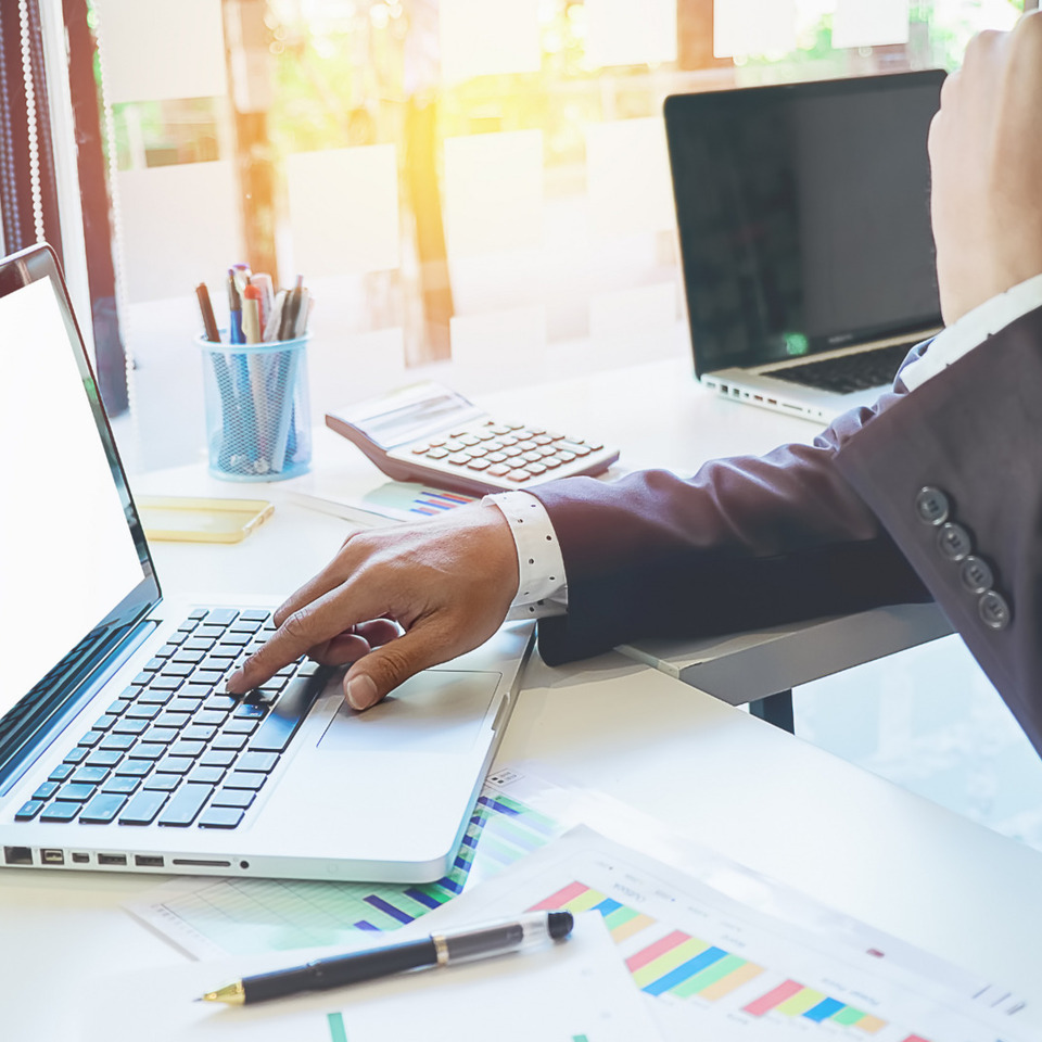 Person in suit working on a laptop at a desk with charts and calculator in a bright office space