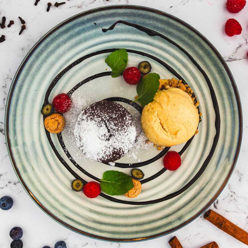 Chocolate cake dessert with a scoop of vanilla ice cream, raspberries, mint leaves, and chocolate drizzle on a stylish plate.