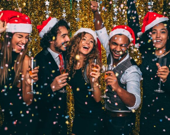 Group of guests in Santa hats toasting champagne amid confetti and gold tinsel backdrop at festive hotel party.