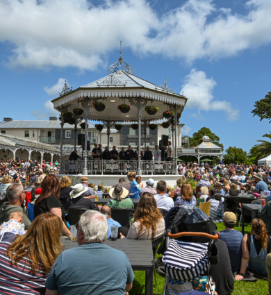 Outdoor bandstand concert on a sunny lawn, Victorian gazebo stage with musicians, crowded audience enjoying festival