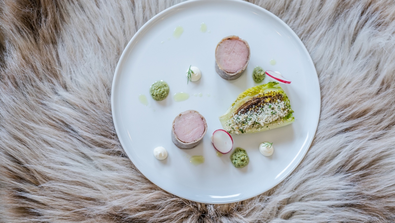 Wykwintne danie: plastry polędwicy owiniętej, grillowana sałata rzymska z kruszonką, plasterki rzodkiewki i kleksy pesto na białym talerzu