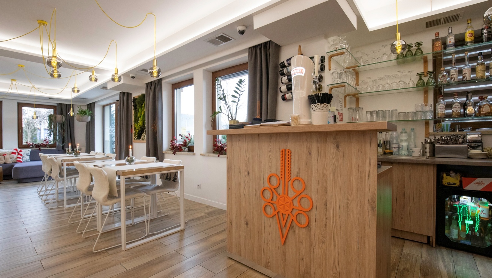 Przytulny hotelowy bar i jadalnia z drewnianą ladą, półkami ze szkłem i alkoholem, białe stoliki, wiszące żarówki i rośliny.