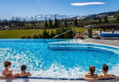 Ogrzewany basen z jacuzzi na świeżym powietrzu, goście relaksują się przy krawędzi, w tle ośnieżone szczyty i zielony teren.