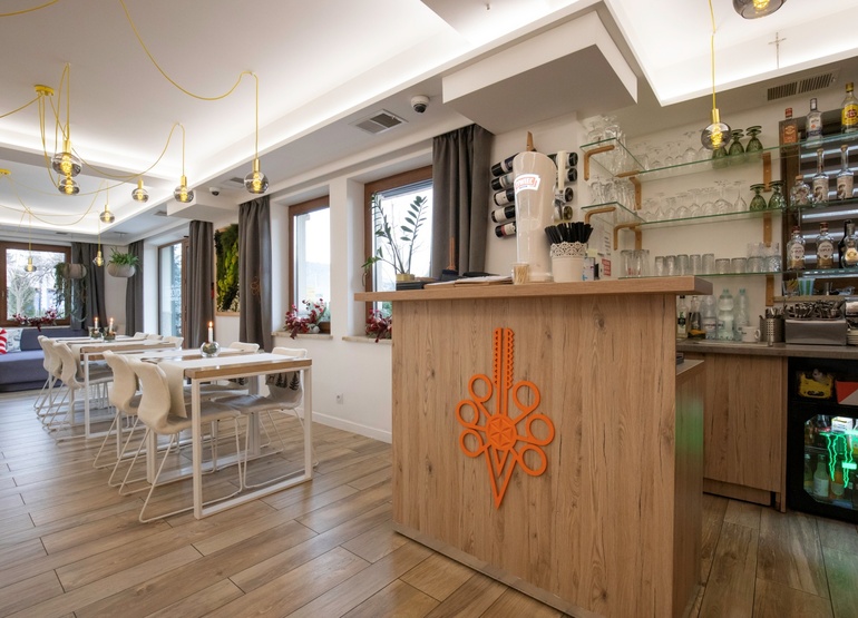 Przytulny hotelowy bar i jadalnia z drewnianą ladą, półkami ze szkłem i alkoholem, białe stoliki, wiszące żarówki i rośliny.
