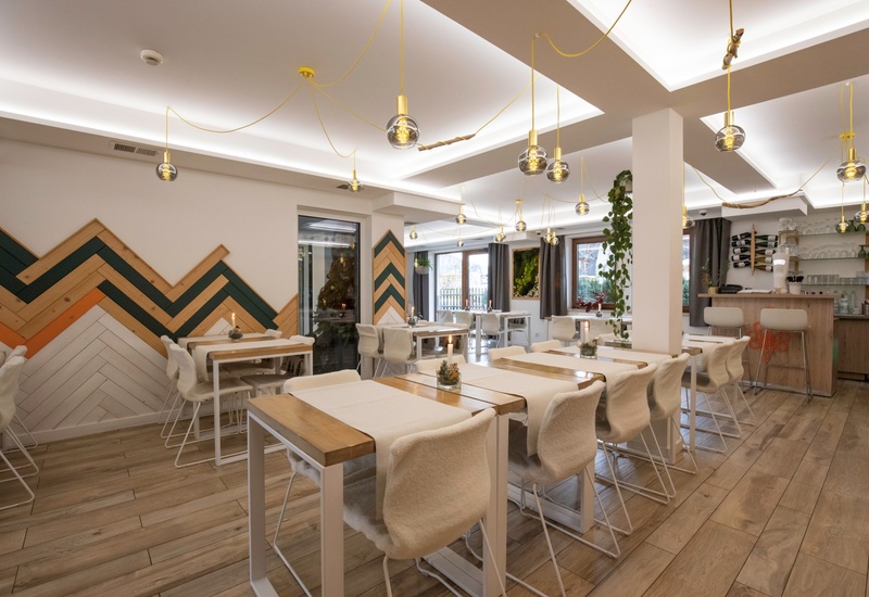 Przytulna restauracja z drewnianymi stołami, futrzanymi krzesłami, szklanymi lampami na żółtych przewodach, barem, oknami i dekoracyjną ścianą