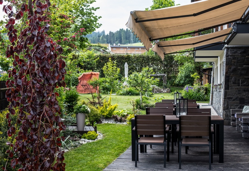 Taras restauracyjny z zacienionymi markizami, stolikami i lampionami, otoczony bujnym ogrodem i widokiem na las.