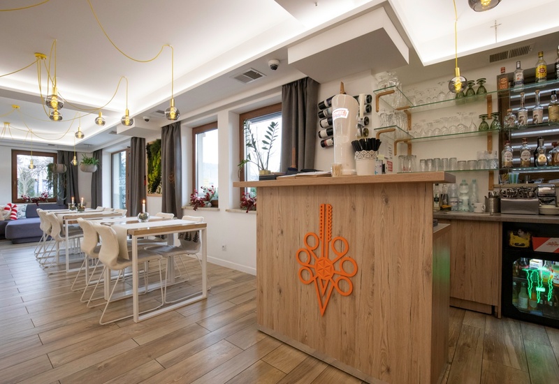 Przytulny hotelowy bar i jadalnia z drewnianą ladą, półkami ze szkłem i alkoholem, białe stoliki, wiszące żarówki i rośliny.
