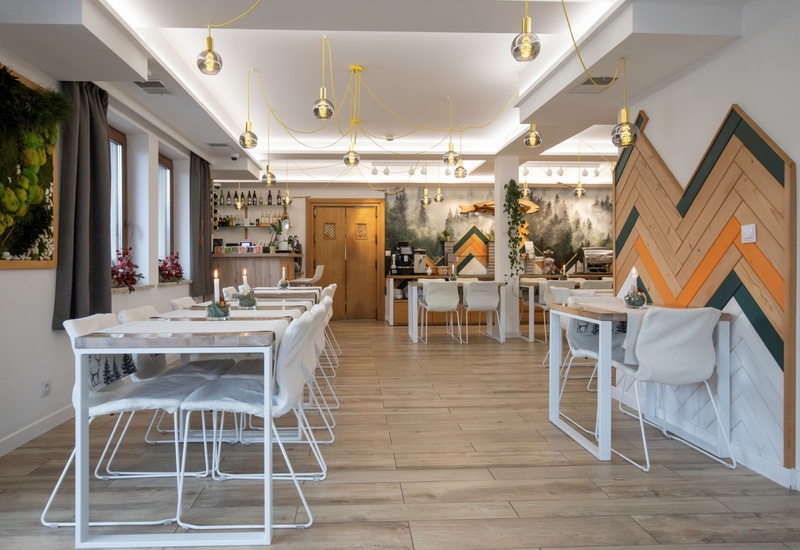 Przytulna restauracja hotelowa z jasnymi drewnianymi stołami, miękkimi narzutami na krzesłach, szklanymi lampami, muralem lasu i barem.