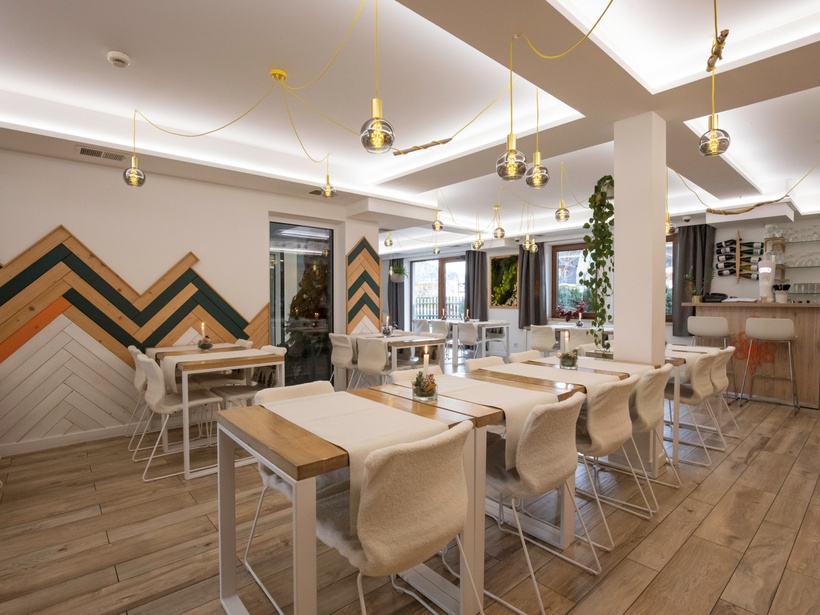 Przytulna restauracja z drewnianymi stołami, futrzanymi krzesłami, szklanymi lampami na żółtych przewodach, barem, oknami i dekoracyjną ścianą
