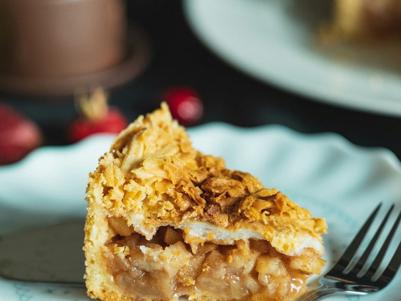 Kawałek szarlotki z chrupiącą kruszonką na pastelowym talerzu, widelec obok, kawa i owoce w tle