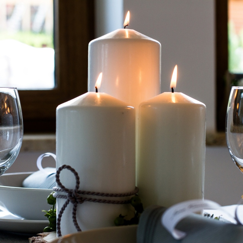 Trzy zapalone białe świeczki typu pień jako dekoracja stołu między kieliszkami. Przytulna restauracja organizująca przyjęcia okolicznościowe.