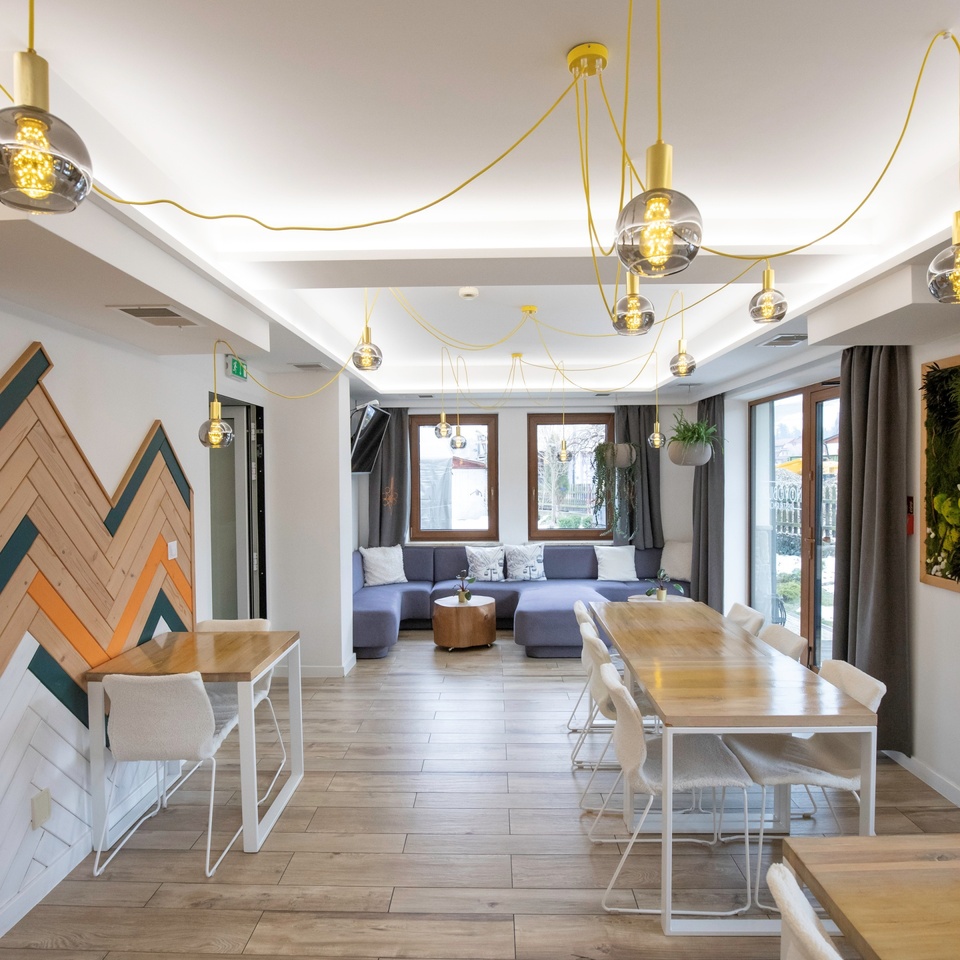 Nowoczesny hotelowy salon z drewnianymi stołami, fioletową sofą, wiszącymi żarówkami na żółtych przewodach i doniczkami