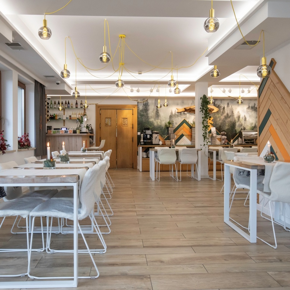 Przytulna restauracja hotelowa z jasnymi drewnianymi stołami, miękkimi narzutami na krzesłach, szklanymi lampami, muralem lasu i barem.
