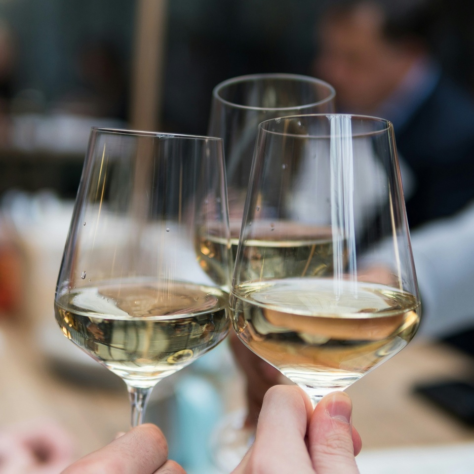 Trzy kieliszki białego wina podczas toastu przy stoliku restauracyjnym, rozmyte sylwetki gości, kameralna atmosfera.