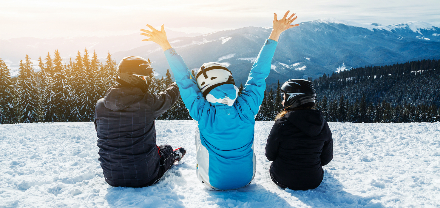 Three helmeted skiers sit in snow on a mountain ski resort, one raises arms toward sunlit alpine peaks and pine slopes