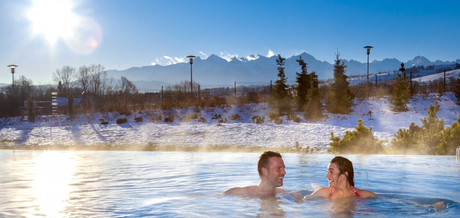 Couple relaxing in a heated outdoor pool with steam, snowy shores and a dramatic mountain vista at sunrise