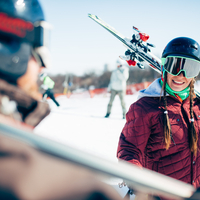 Smiling skier in helmet and goggles carrying skis on shoulder at a sunny resort slope, ready for a run