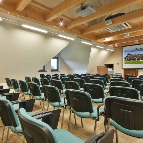 Konferenzraum mit sichtbaren Holzträgern, Reihen mit türkisfarbenen Stühlen, Projektor und Leinwand, Rednerpult und Klimaanlage