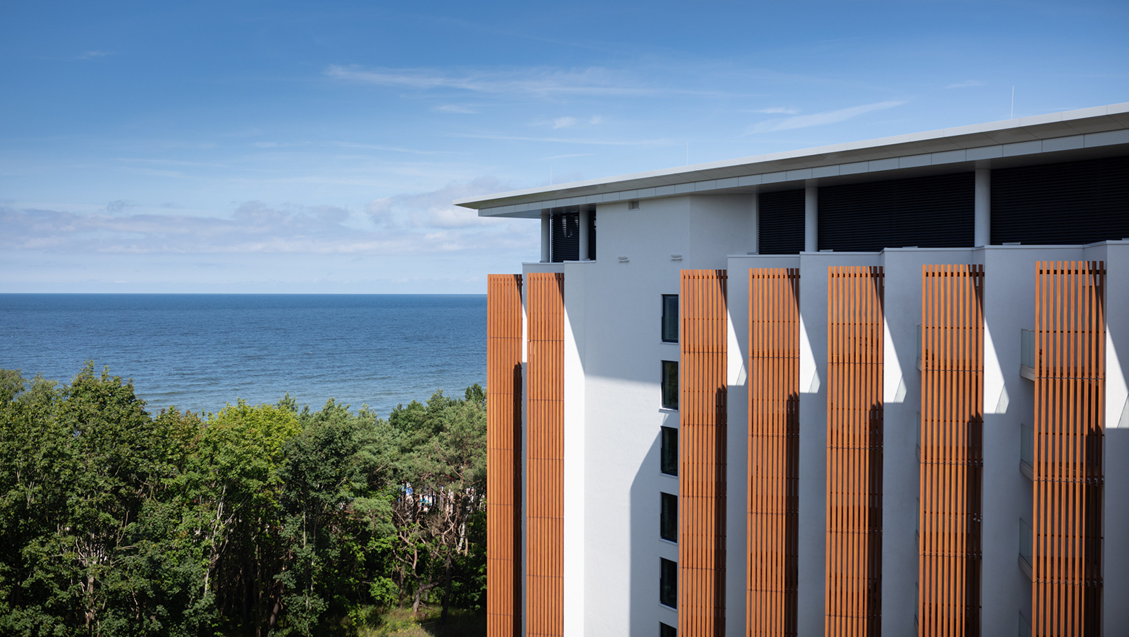 Nowoczesny hotel przy morzu z drewnianymi panelami na elewacji, widok na las i bezkresny błękit oceanu.