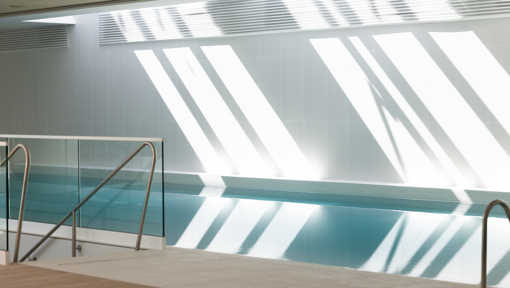 Nowoczesny, jasny basen kryty z metalowymi poręczami i szklaną barierką, naturalne światło tworzy ciekawe cienie