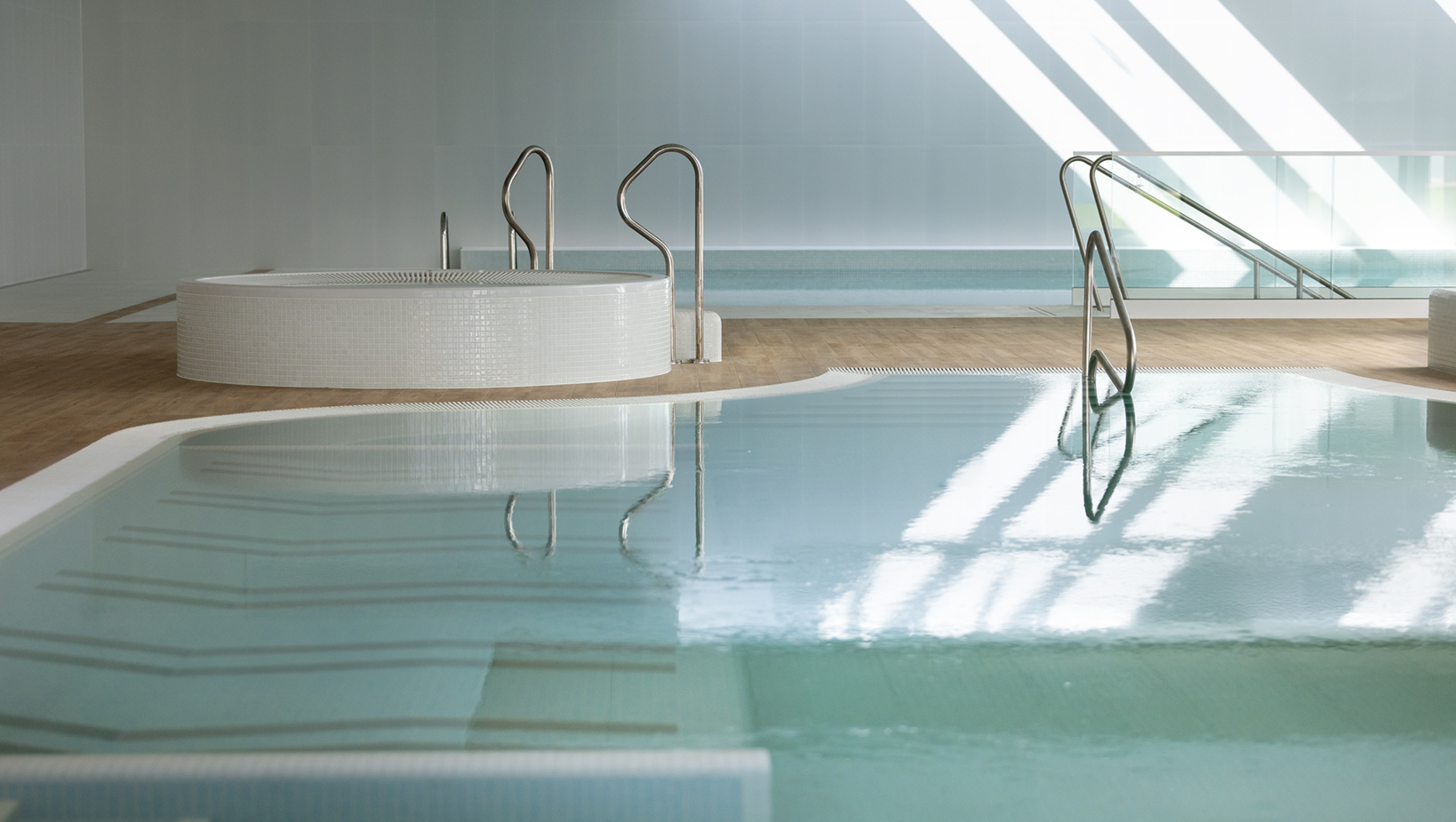 Nowoczesny, jasny basen w hotelu z białymi kafelkami, schodami i okrągłym jacuzzi na drewnianej podłodze.