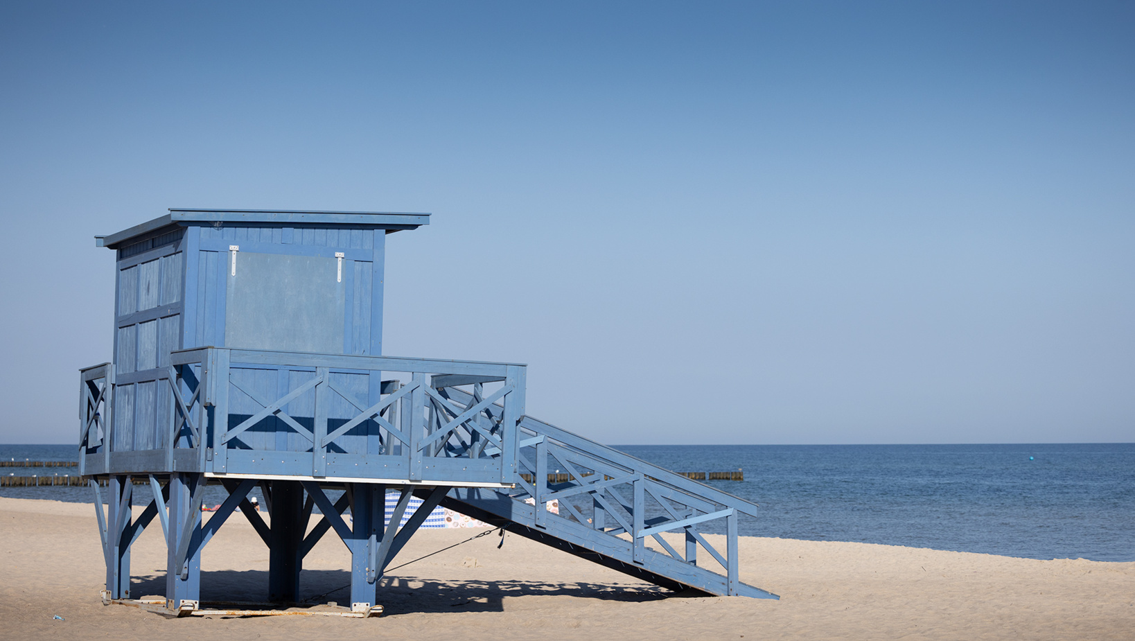 Niebieska drewniana wieża ratownika na piaszczystej plaży z widokiem na spokojne morze i klarowne niebo.