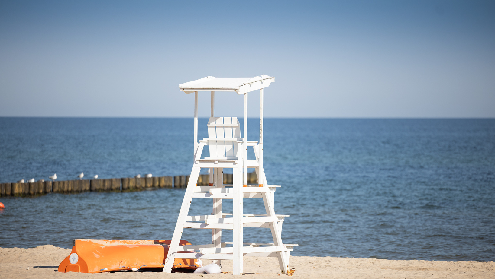 Biała drewniana wieża ratownika na piaszczystej plaży z widokiem na spokojne morze i drewniany falochron.