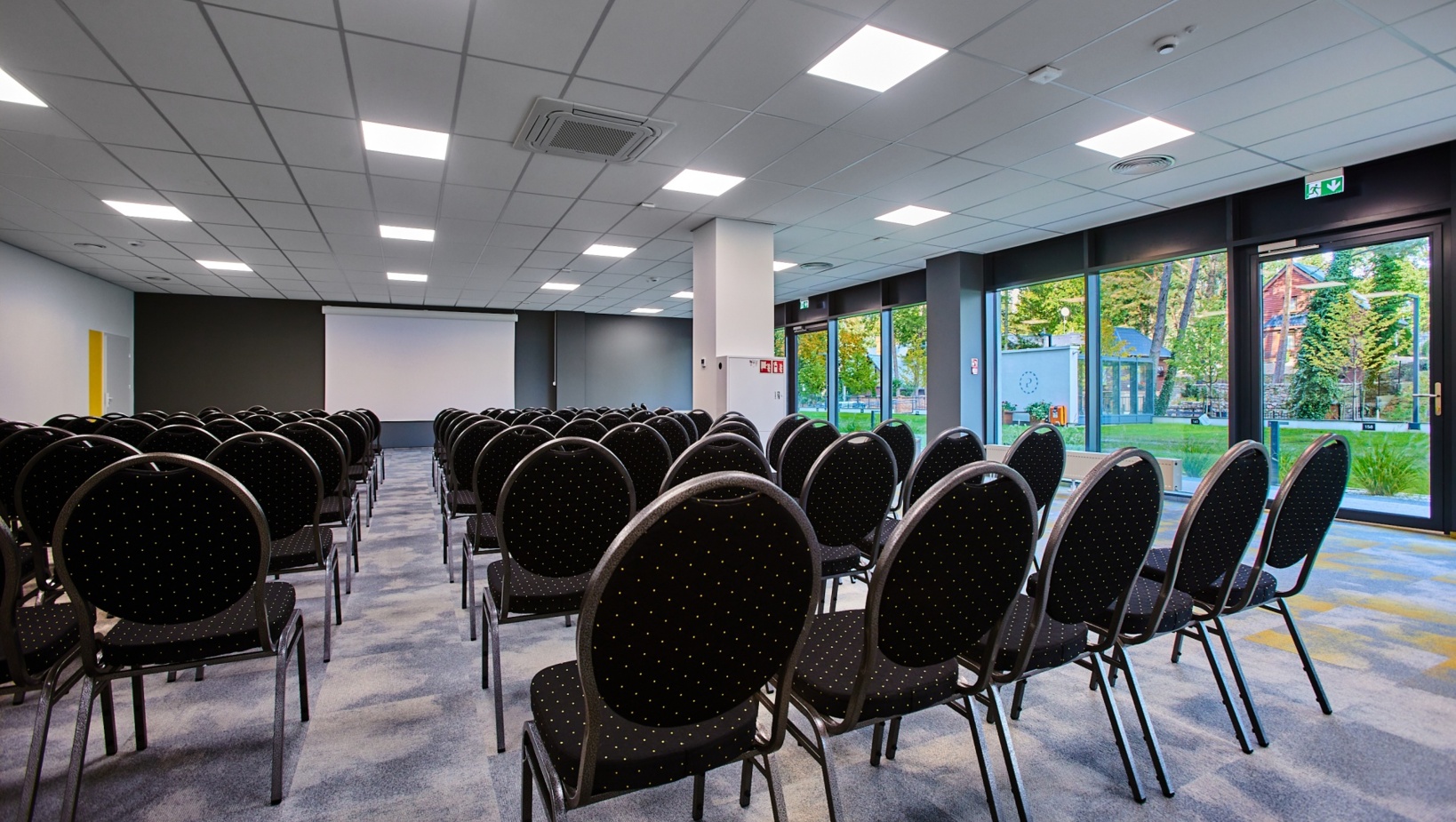 Nowoczesna sala konferencyjna z rzędem czarnych krzeseł, projektorem i dużymi oknami z widokiem na ogród.
