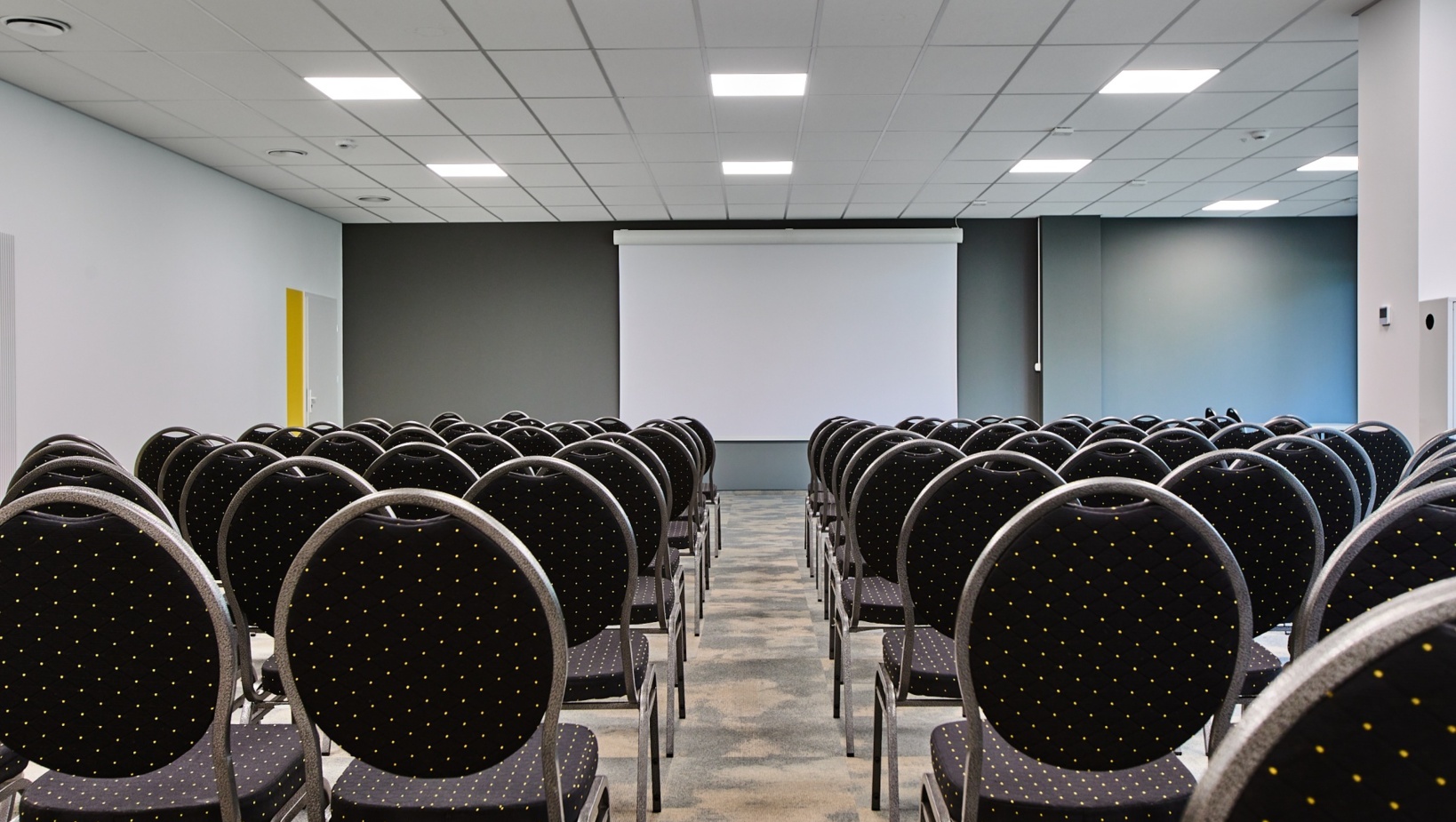 Nowoczesna sala konferencyjna z rzędami czarnych krzeseł i dużym ekranem projekcyjnym na szarej ścianie