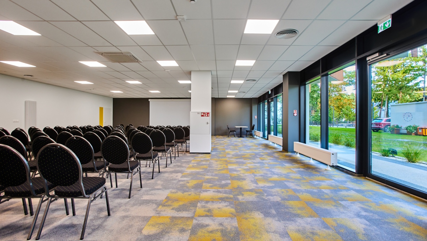 Nowoczesna sala konferencyjna z rzędem krzeseł i dużymi oknami z widokiem na zieleń na zewnątrz