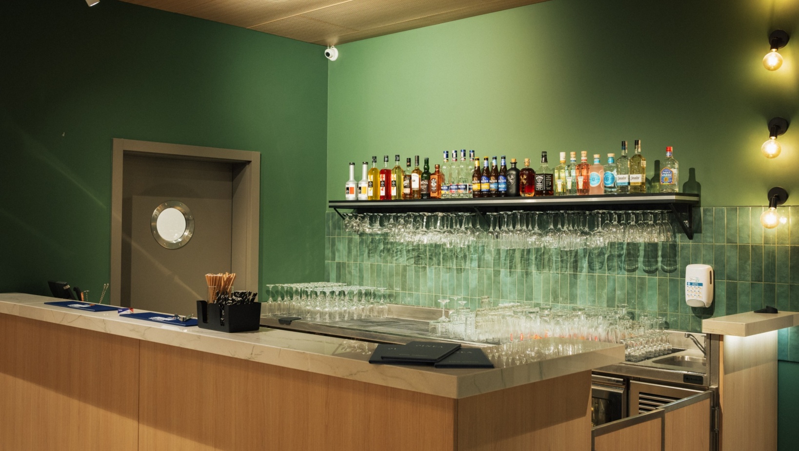 Nowoczesny hotelowy bar z zielonymi ścianami, drewnianym blatem i półką z alkoholem oraz kieliszkami.