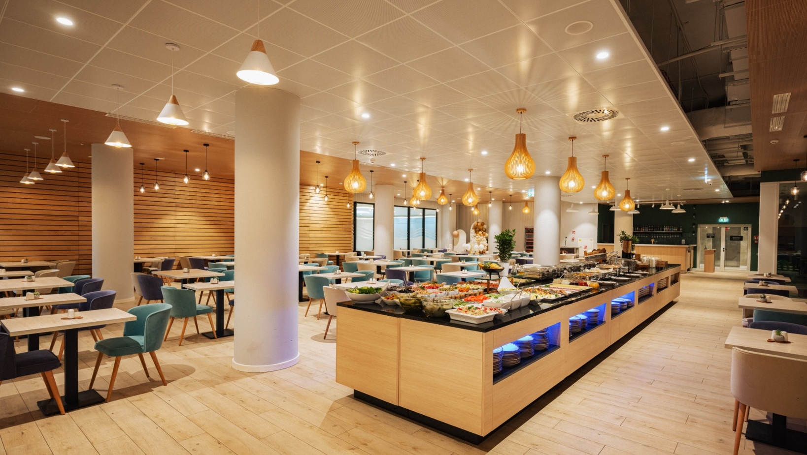 Nowoczesna hotelowa jadalnia z bufetem pełnym świeżych potraw i eleganckimi, kolorowymi krzesłami przy drewnianych stołach.