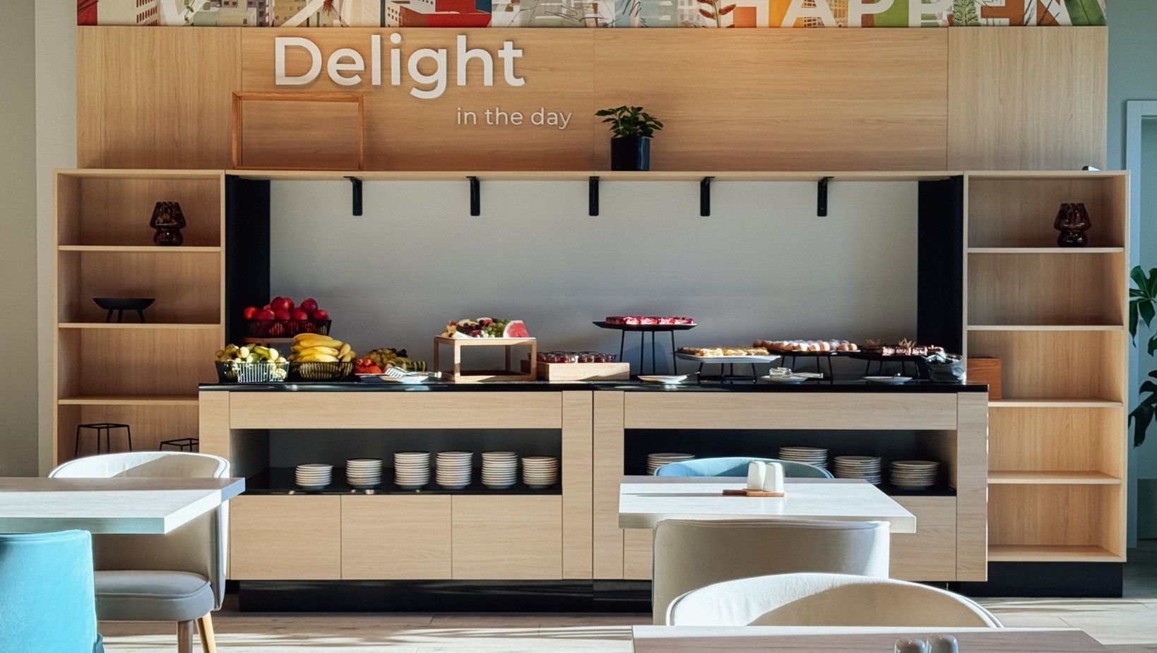 Jasna jadalnia z bufetem oferującym świeże owoce i słodkości, nowoczesne meble i dekoracje ścienne z napisem Delight