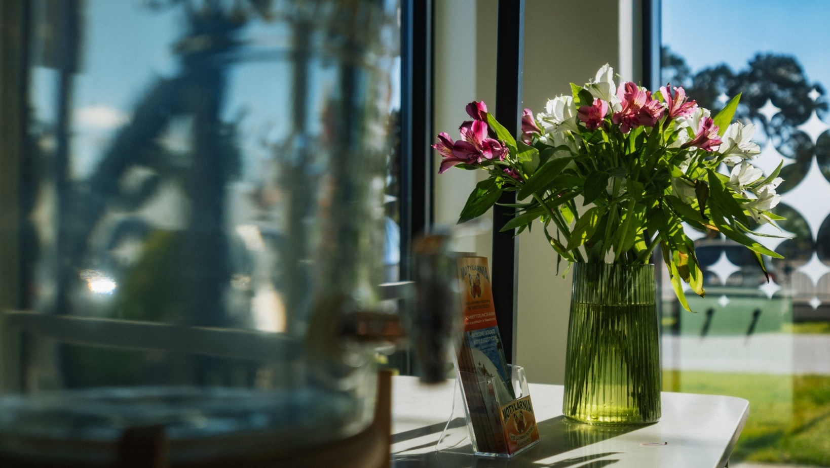 Wazon z różowymi i białymi kwiatami ustawiony na białym blacie przy oknie hotelowym z widokiem na zielony ogród